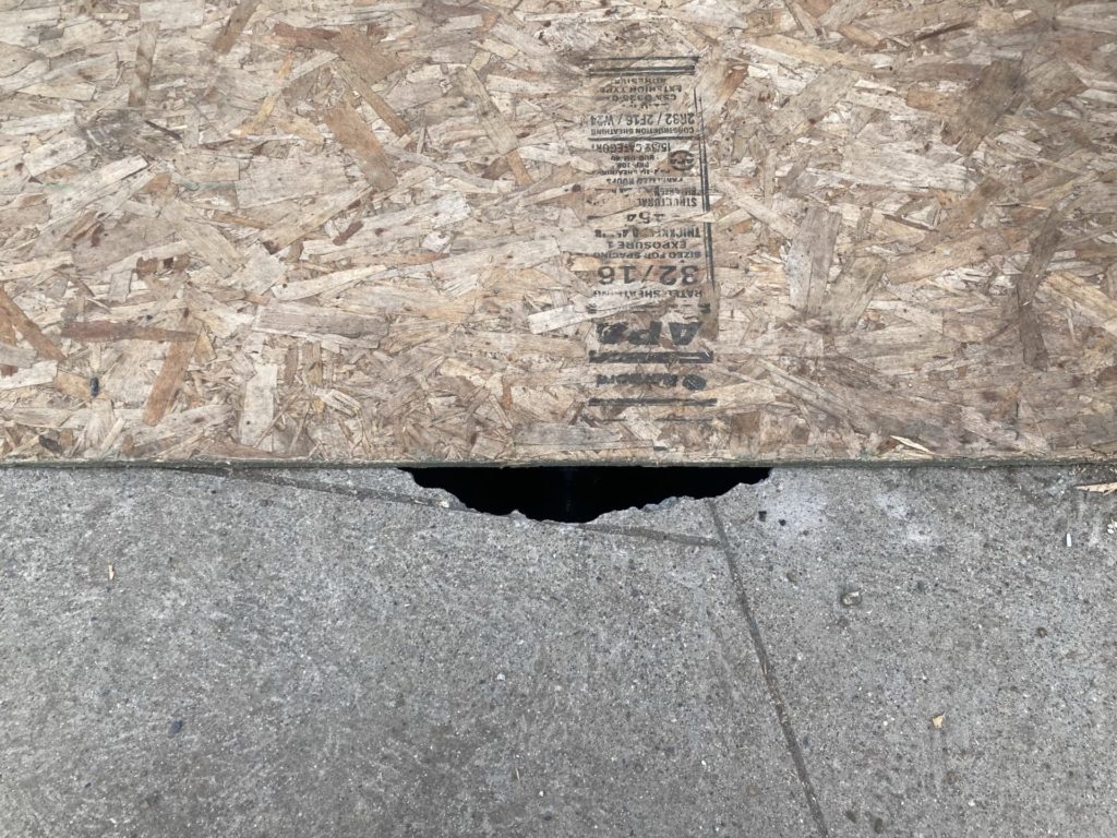 Hole in sidewalk partially covered by plywood