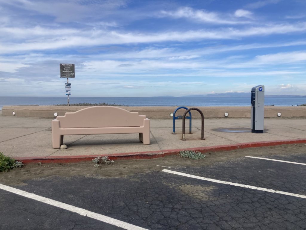 In the foreground are white parking stripes on black pavement. A beige bench, bike rack and parking lot payment machine are on the sidewalk with ocean behind and blue sky streaked with clouds