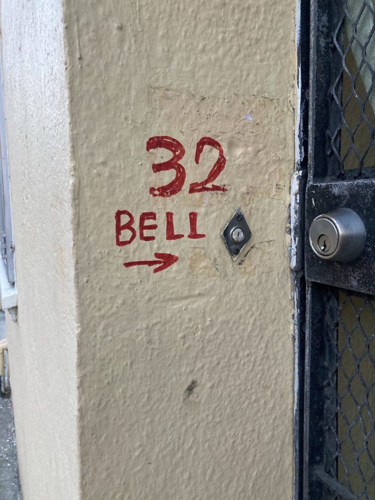 Beige wall with “32 BELL” and an arrow painted in red. The arrow points to a doorbell. A doorway gate lock is next to the doorbell. 