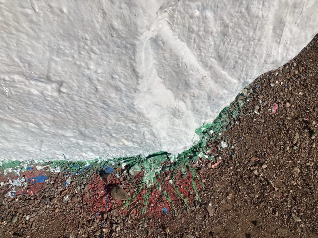 Large white painted rock in top half of frame and brown ground in bottom half. The ground is splattered with different colored paints.