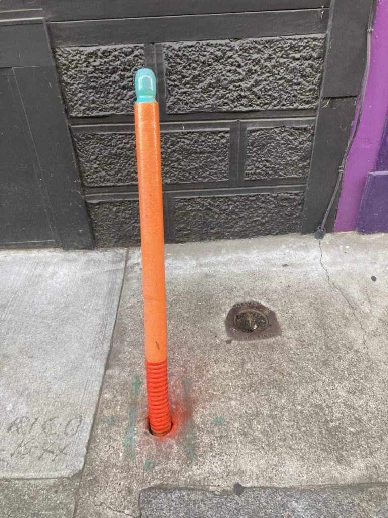 Thin bollard painted light blue, orange and red, against a brick wall painted dark gray. 