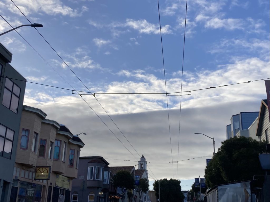 Looking down the street with storefronts, street lamps and bus wires, the sky is blue with some fluffy clouds and a solid wave of clouds.
