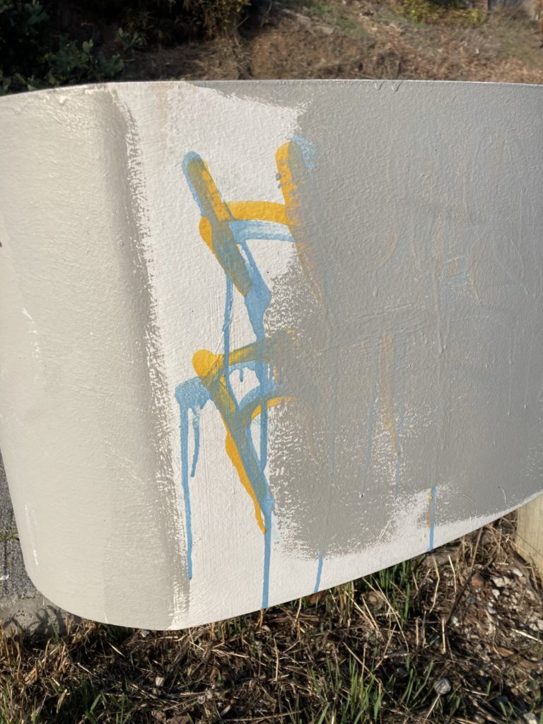 Close up of a guard rail with a drippy yellow and green graffiti tag mostly painted over with gray paint. 