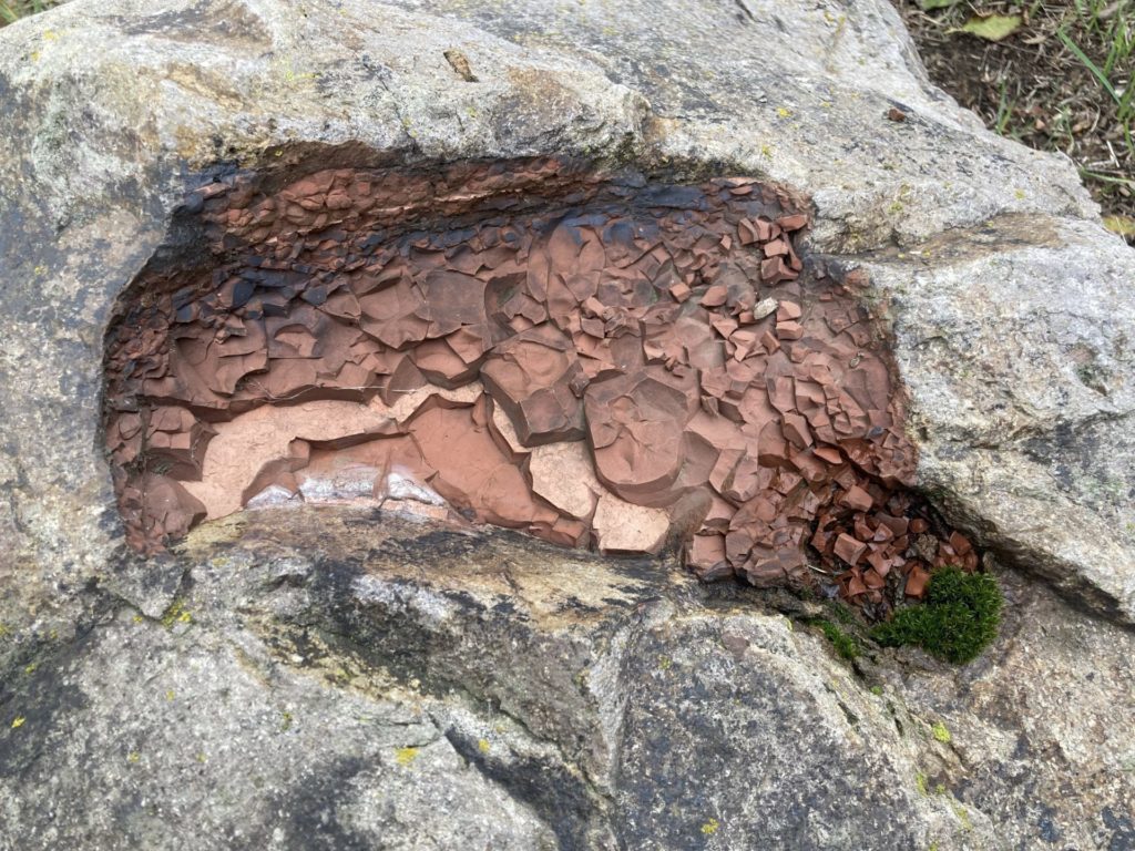 Gray boulder with worn spot showing crumbly, jagged red rock. Some moss grows in the lower right corner of the red rock. 