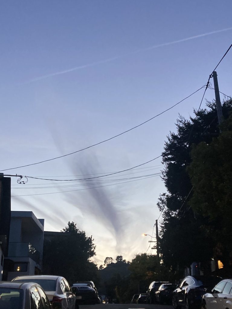 Looking down a street with cars parked on either side. The dusky blue sky is clear except for a vertical whisp of gray cloud. Power lines arch over the street. 