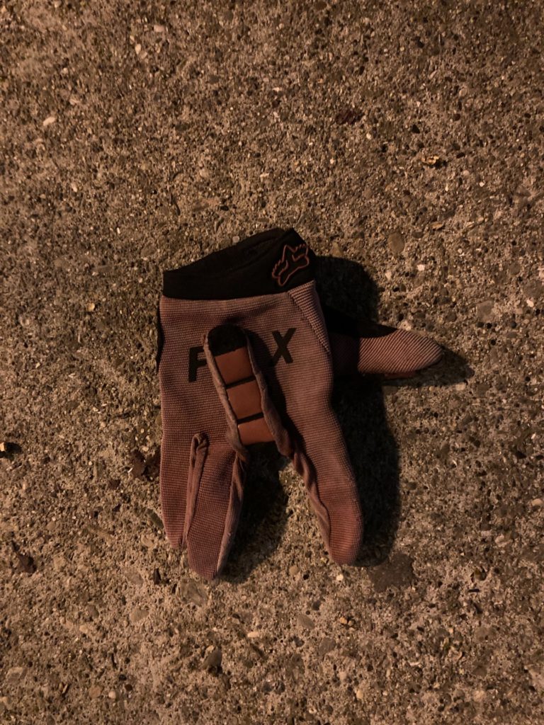 Brown glove on the sidewalk at night, illuminated by a street lamp. The fingers face down except the middle finger, folded up toward the back of the palm.