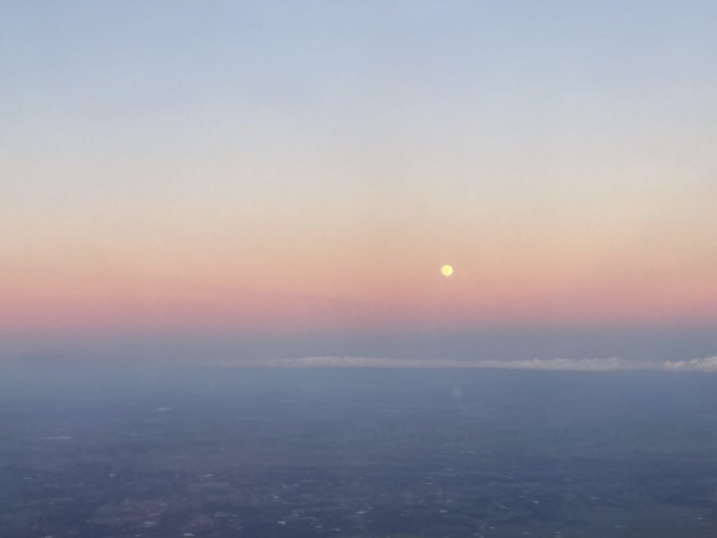 View from plane of light blue sky and dark blue ground, bisected by a pink band of light from sunset. The full moon is visible in the pink band. 