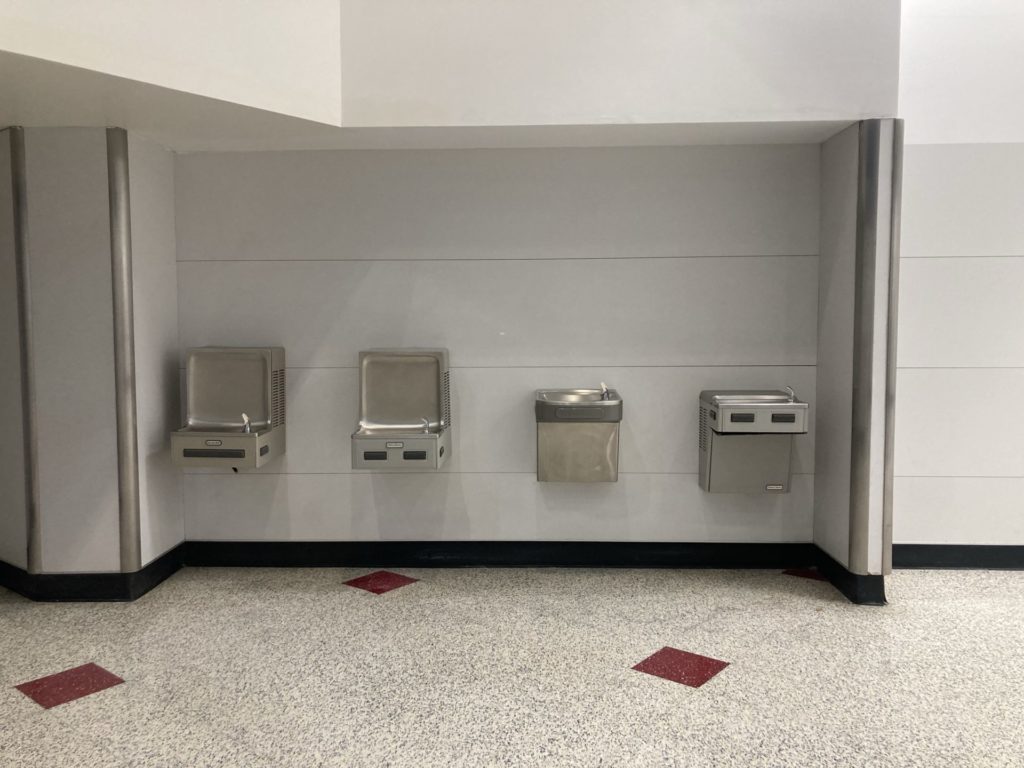 Wall with four different water fountains, each one different. The floor is white speckled tile with occasional red diamond tile. 