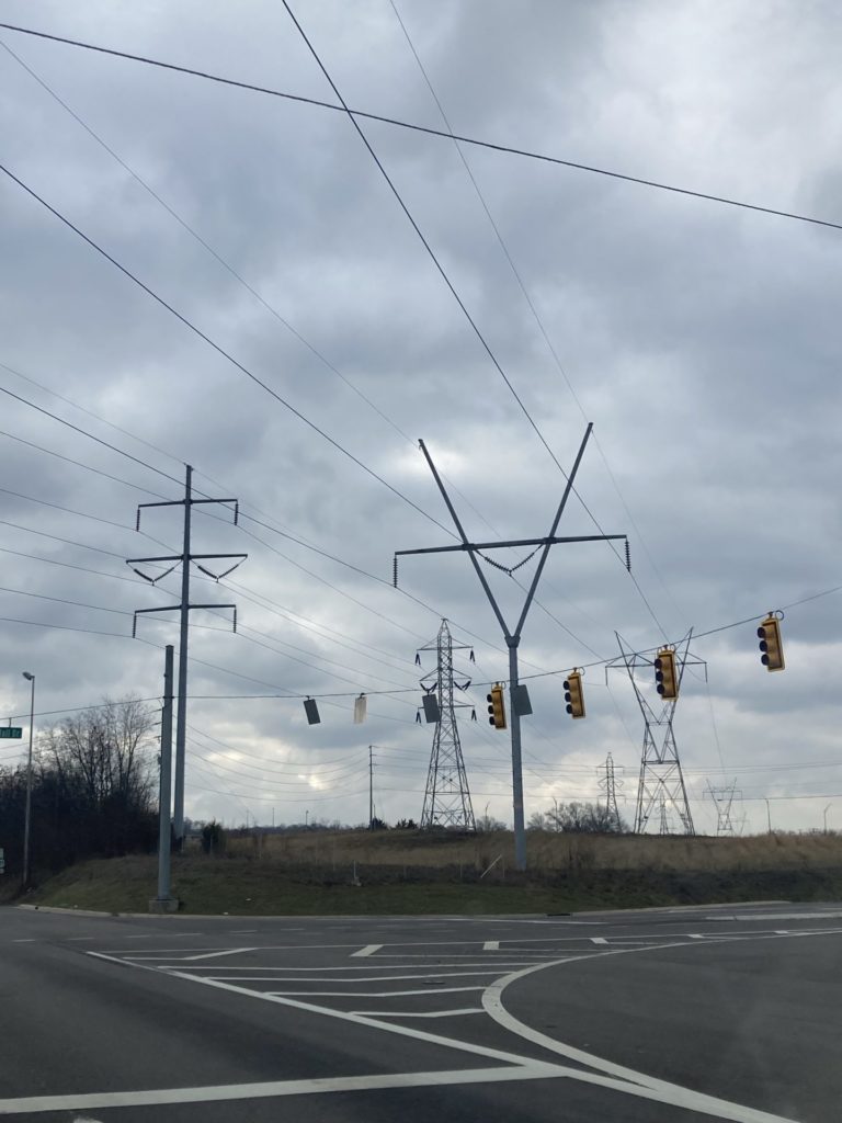 Four different shaped power line poles against a gray cloudy sky. Traffic stripes are visible on the road in the foreground. 