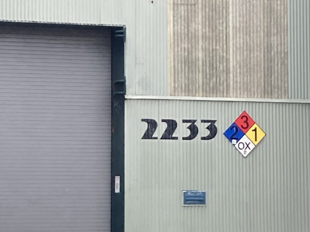 Industrial building wall clad in vertical light green and gray corrugated metal. A garage door is on the left, in the middle are the numbers 2233 written in a bold art deco font. To the right is a diamond shaped material safety sign. A mail slot is below the numbers