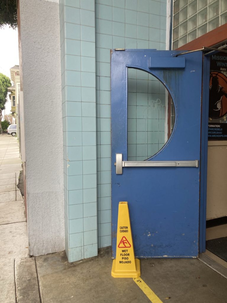 Beige stucco wall next to aqua blue tile wall. A door painted dark blue with a half circle window is propped open against the blue tile with a yellow “wet floor” cone