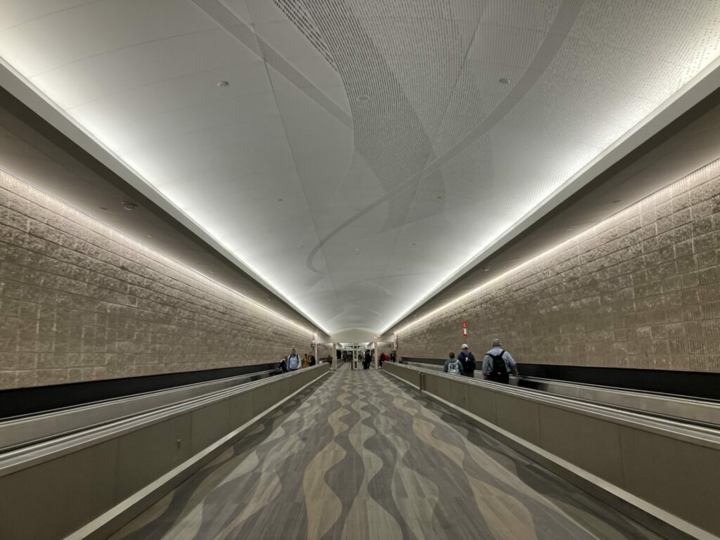 Long beige hallway with moving sidewalks on either side, wavy patterned carpet between them, and gently lit ceiling
