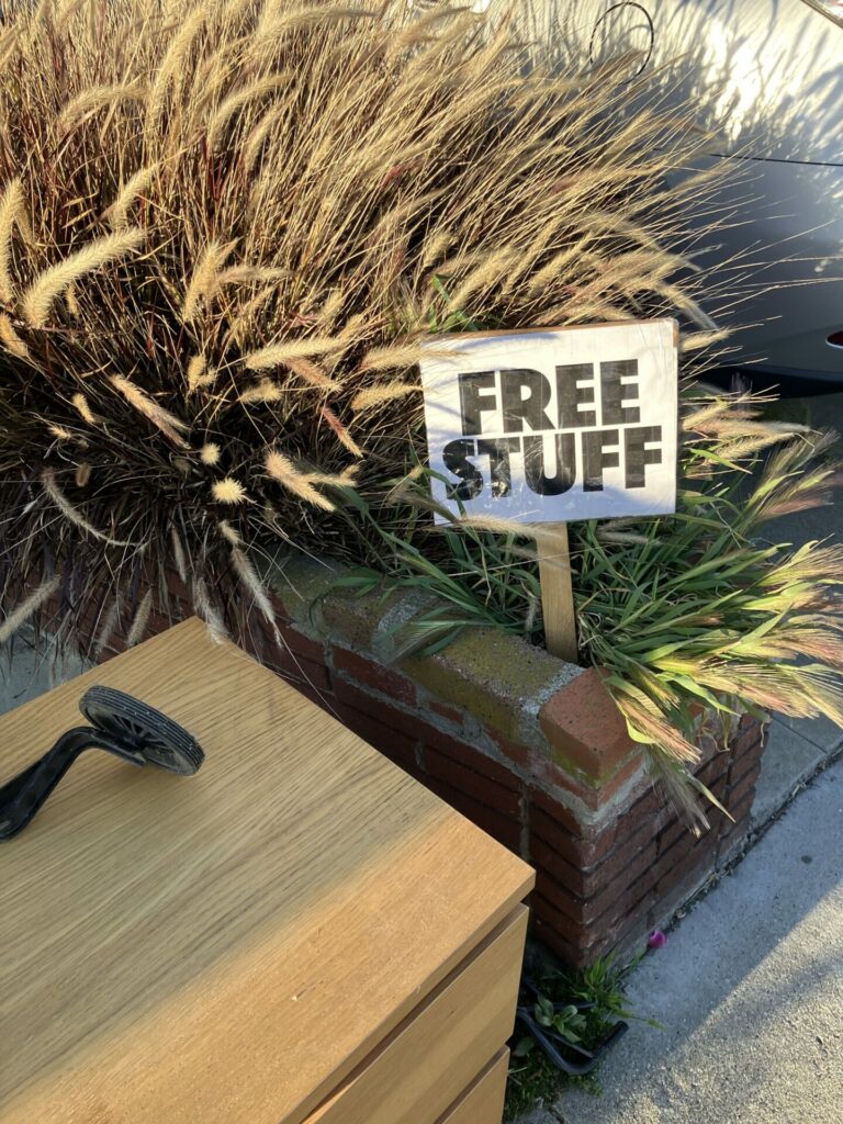Computer print out sign “FREE STUFF” stuck in a planter with wispy white grass, next to a light brown cabinet with a training wheel on top of it. 