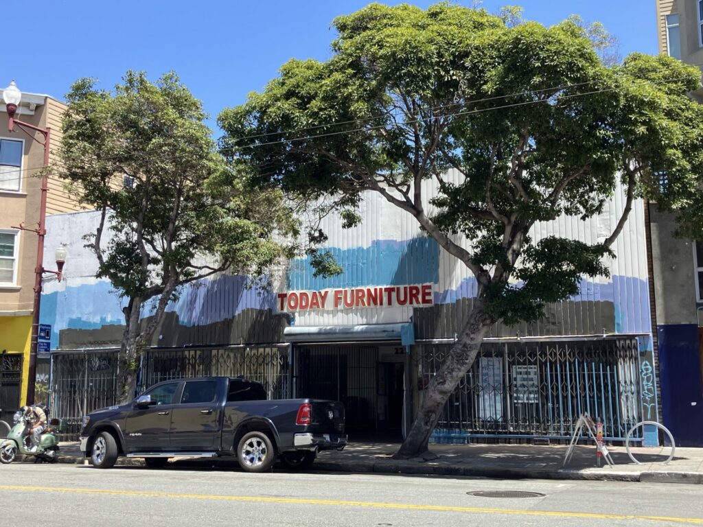 Storefront with red letter sign, “TODAY FURNITURE” and lots of random paint in shades of blue and black, presumably covering graffiti or old signs. Two trees are in front of the building along with a black truck. 