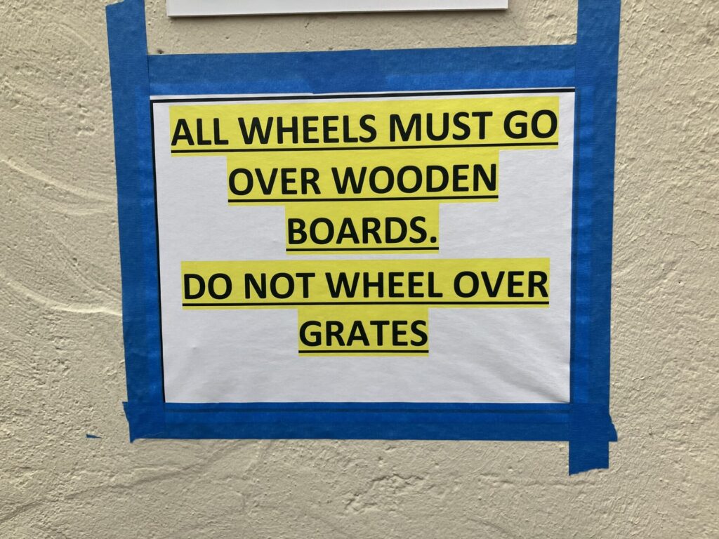 Sign taped to wall reading: ALL WHEELS MUST GO OVER WOODEN BOARDS. DO NOT WHEEL OVER GRATES. The tape is blue and typed letters are bold, all caps, underlined and highlighted. 