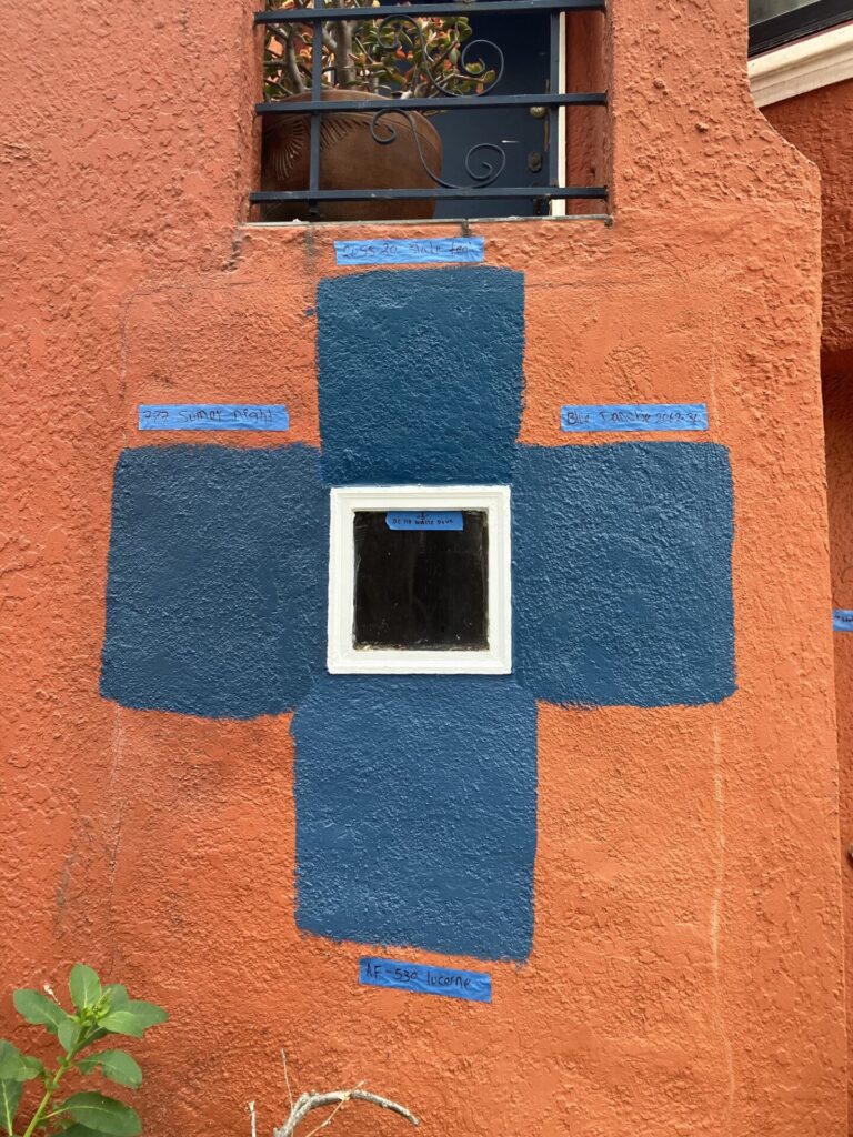 Orange wall with four shades of blue squares painted around a small square window, forming a flower design. The blue shades are labeled with tape. The shades all look pretty much the same. 