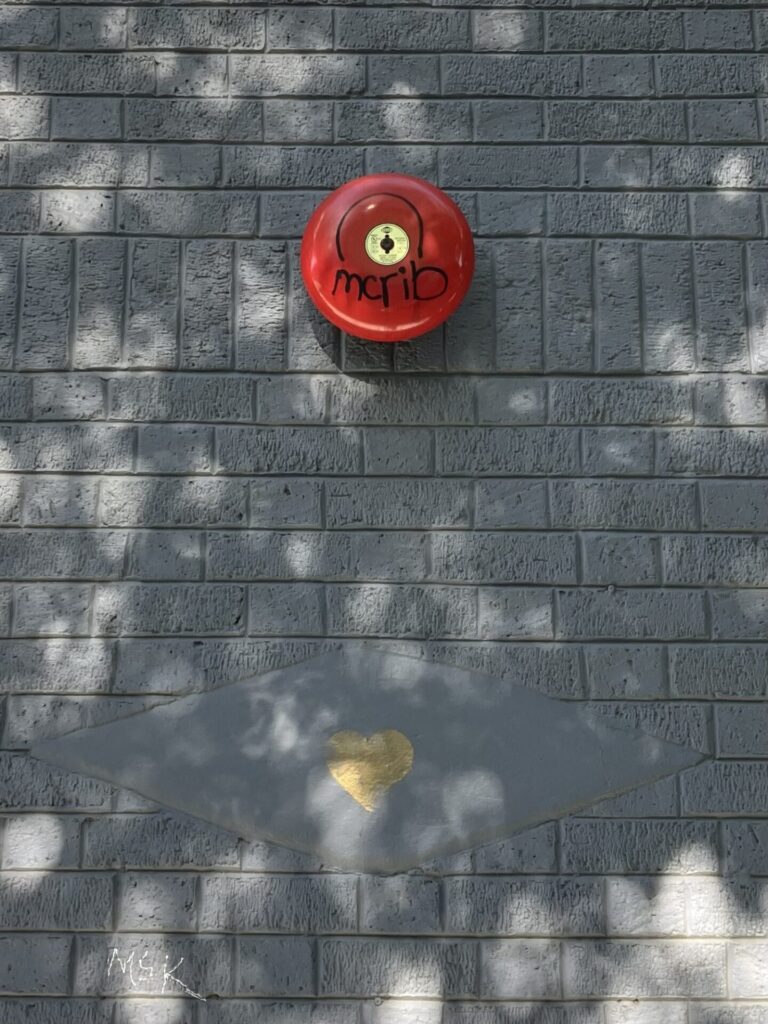 Brick building painted gray with a decorative gray diamond. There is a painted gold heart in the diamond. Above the diamond, a red fire alarm bell with the graffiti “mcrib” in black.