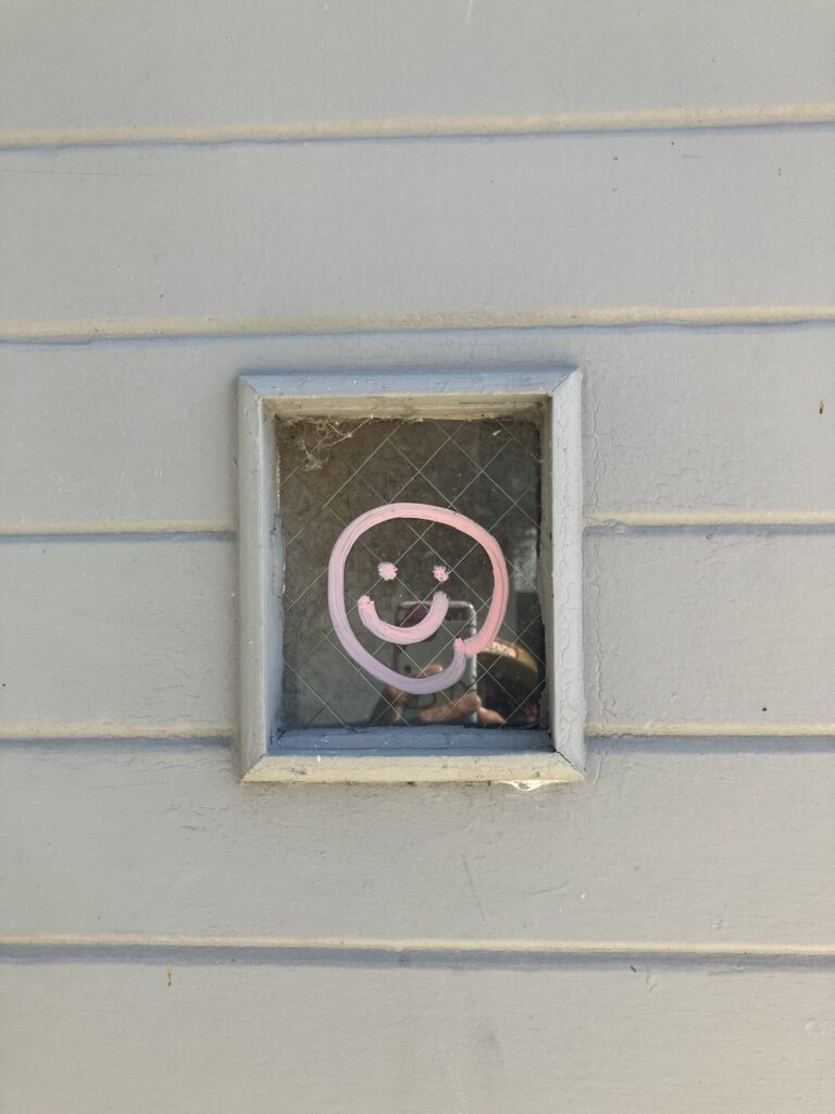 Cream colored wood siding and a small window, on the window a smily face is drawn in pink ink. 
