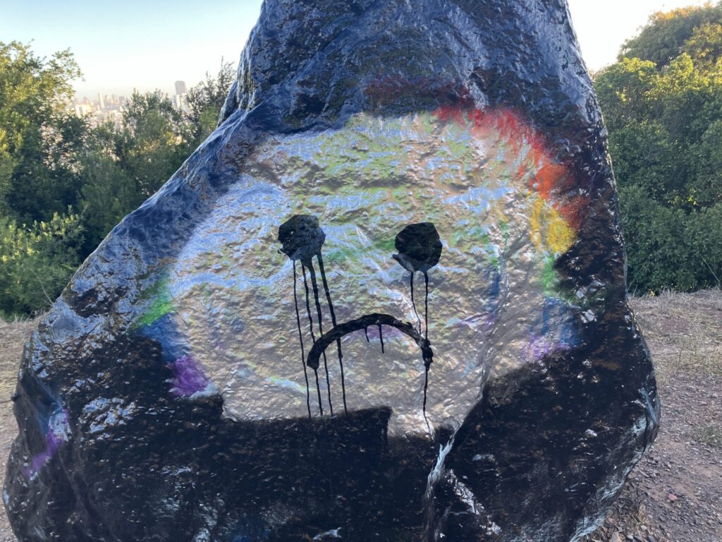 Boulder painted black with silver oval in the middle. A shadow of a rainbow is underneath the silver. A frowny face is painted in drippy paint on the silver oval. 