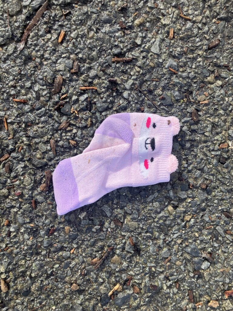 A pink and purple baby sock with a cute bear face, on the sidewalk. 