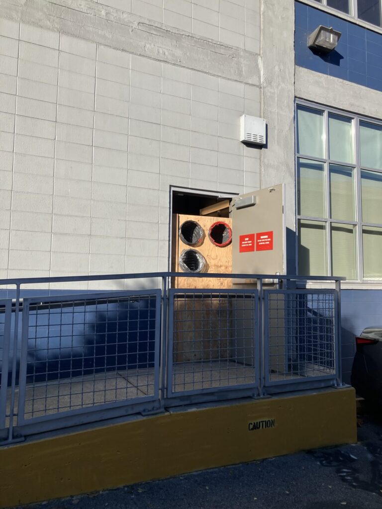 Outside of building with open door. In the doorway is plywood with three large round holes cut near the top, and flexible ducts connected to the holes. 