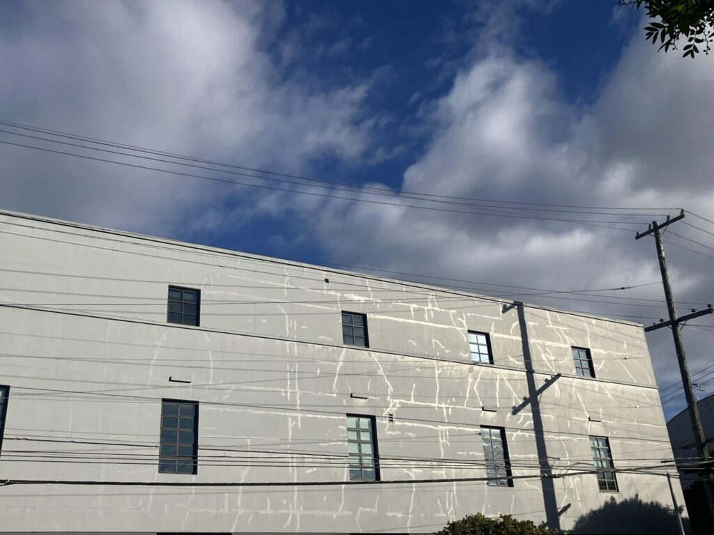 Long side of a gray building in the sun. Lines of stucco repairs or something shine on the right side of the building. The sky is blue with lots of clouds. 
