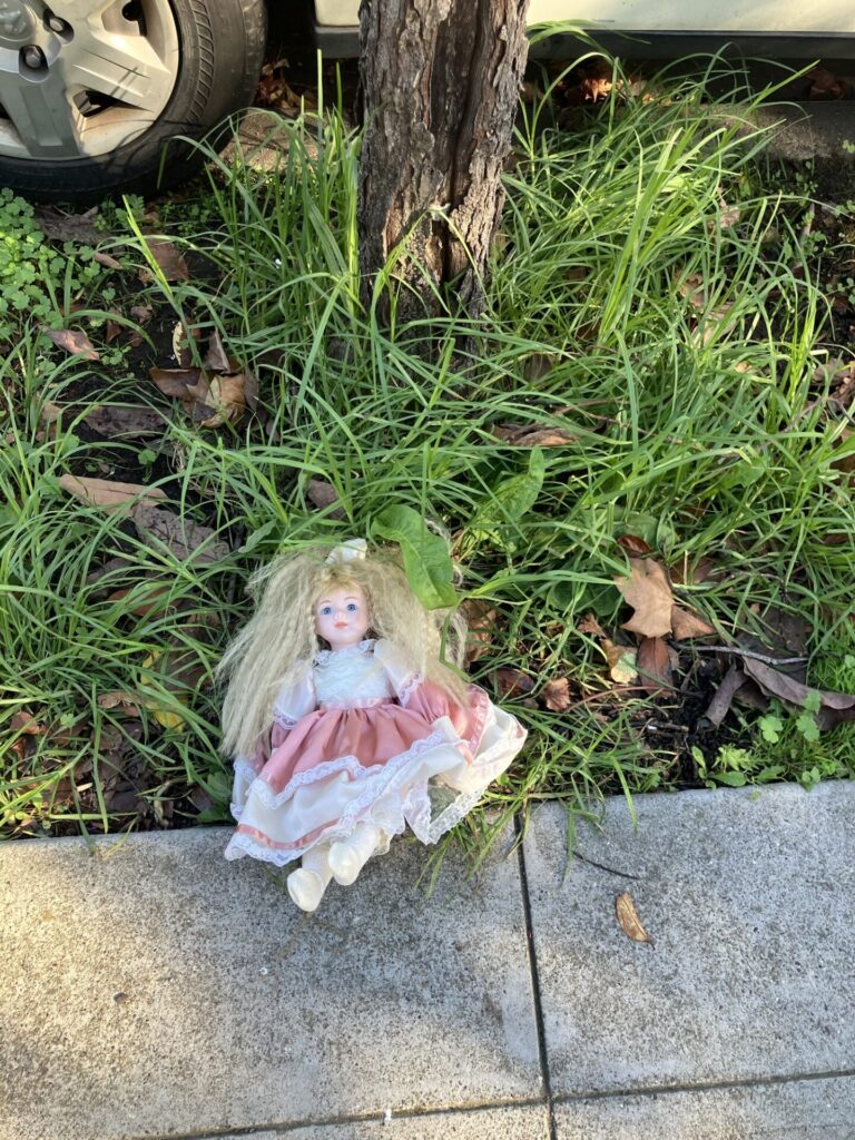 White doll with crimped blonde hair and a white and pink lacy dress in a tree hole in the sidewalk. 