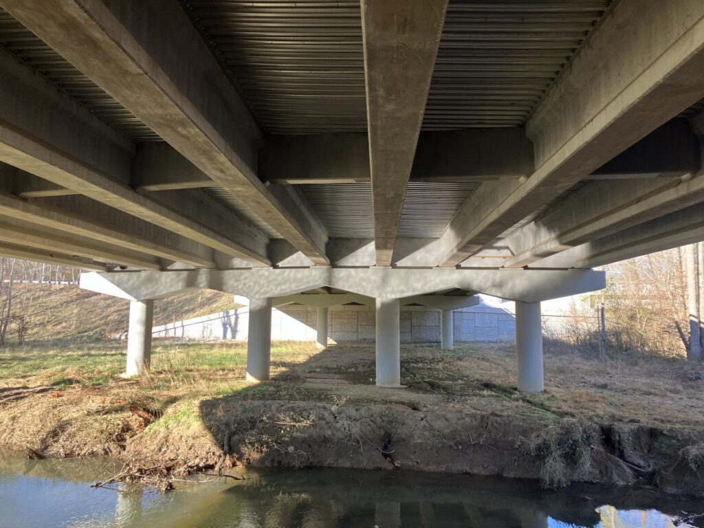 View from under a small bridge, a stream in the bottom of the frame and the underside of the bridge at the top. 