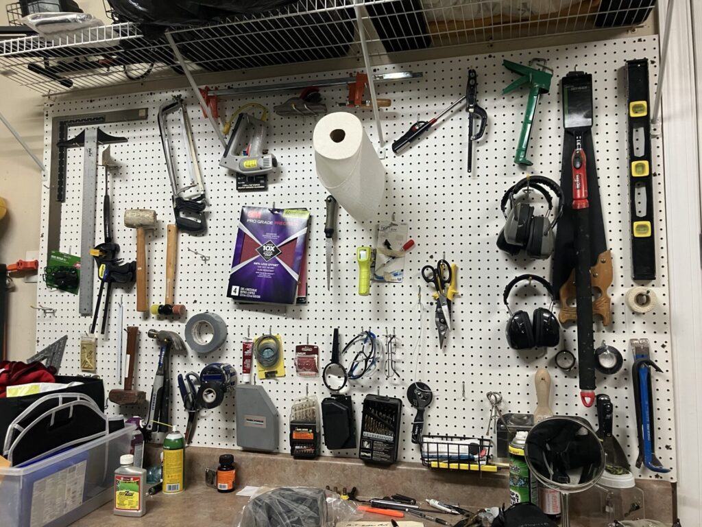 View of wall behind a workbench. The wall is covered in pegboard and hanging from the pegboard are all kinds of tools: level, saw, scissors, hammers, ear protection, roll of paper towels, duct tape, etc. 