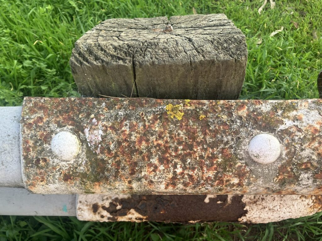 Rusty metal guardrail attached to a wooden post, with green grass in the background. 