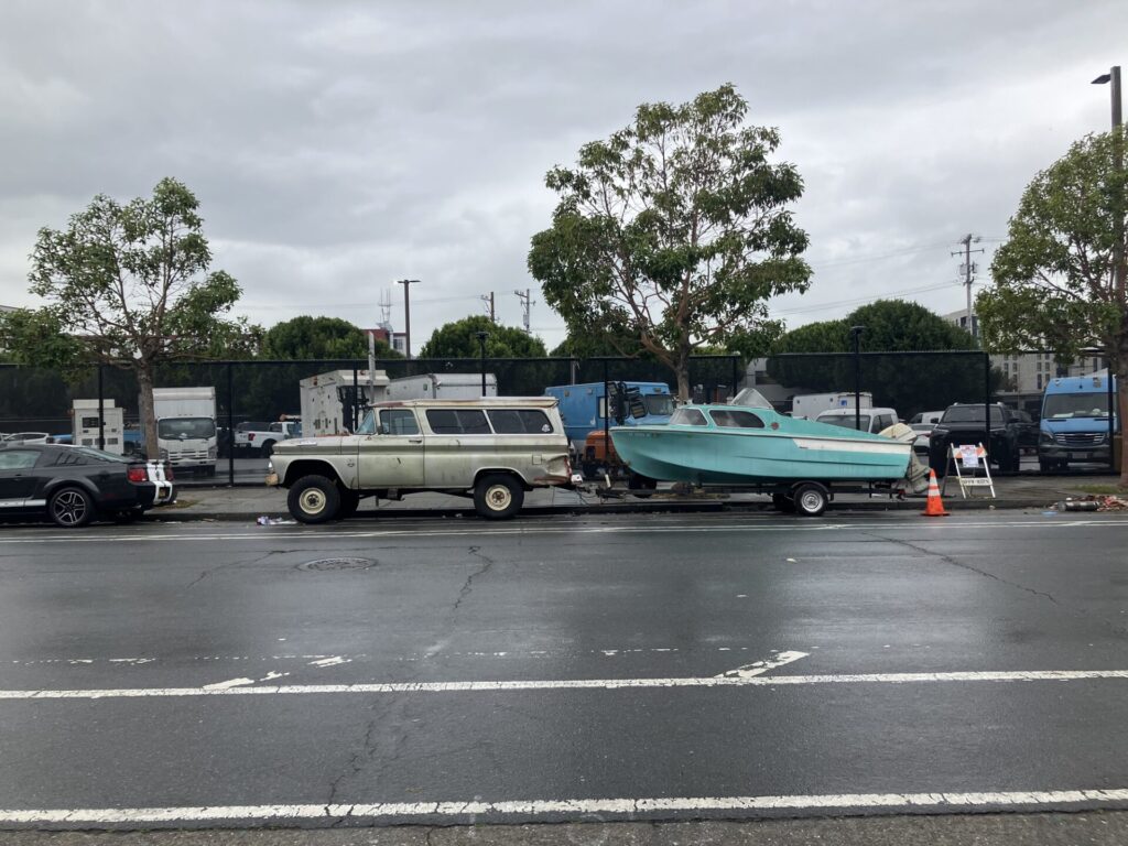 Parked on the street, an old beige truck with bed cover with an old teal boat hitched to it. 