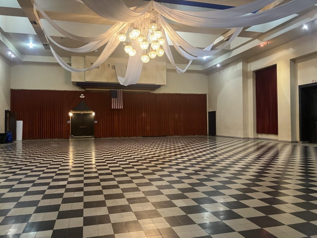 Large empty room with checkerboard floor, red curtains and an american flag along the back wall, and a ball chandelier with curtain streamers along the ceiling. 