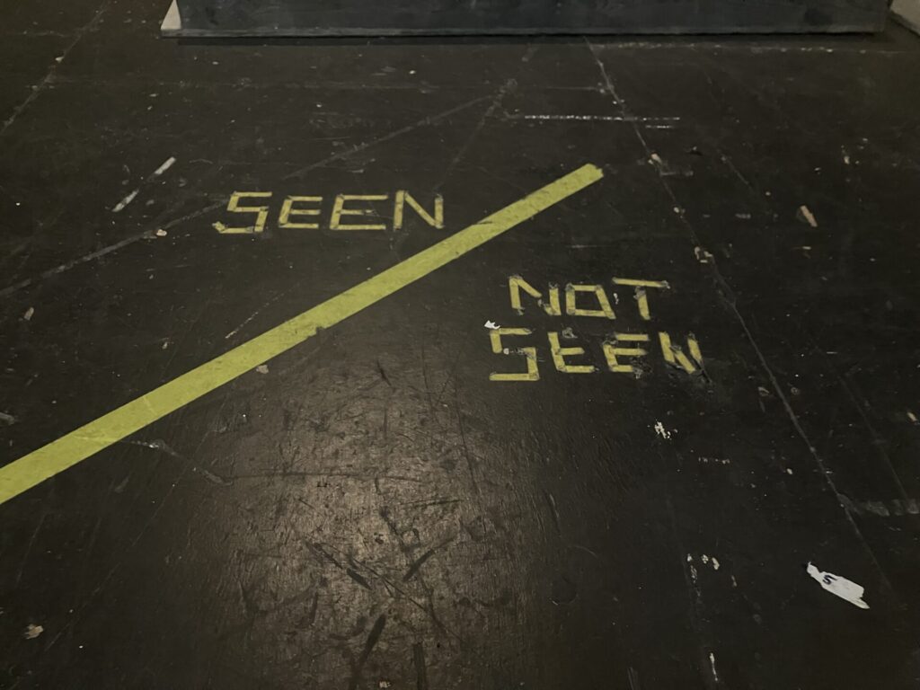 Black floor with words “SEEN / NOT SEEN” written in yellow tape. 