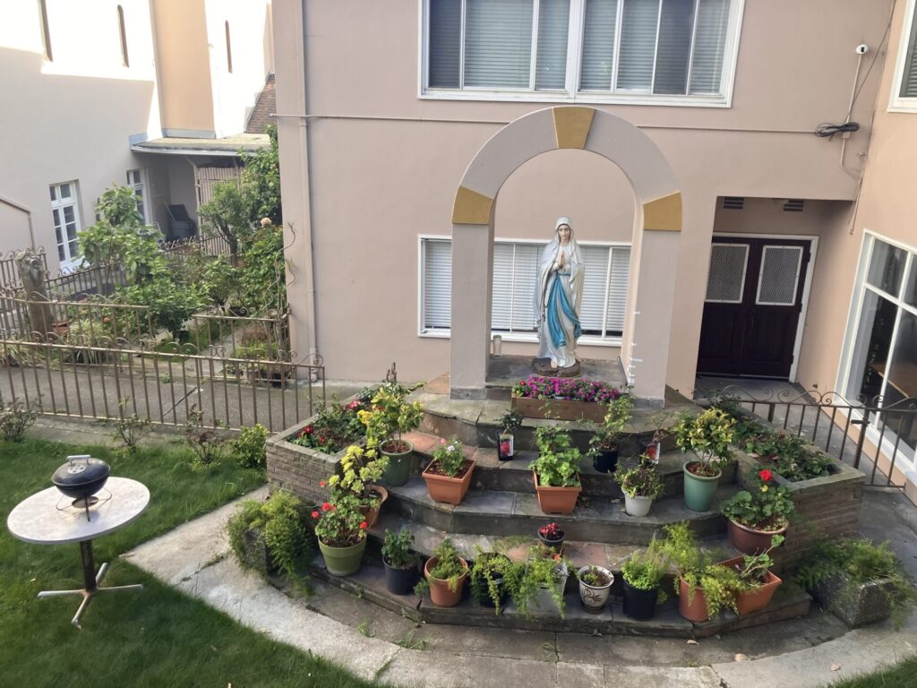 Outdoor shrine to Virgin Mary surrounded by plants. Nearby, a round indoor table with a tabletop grill on it. 