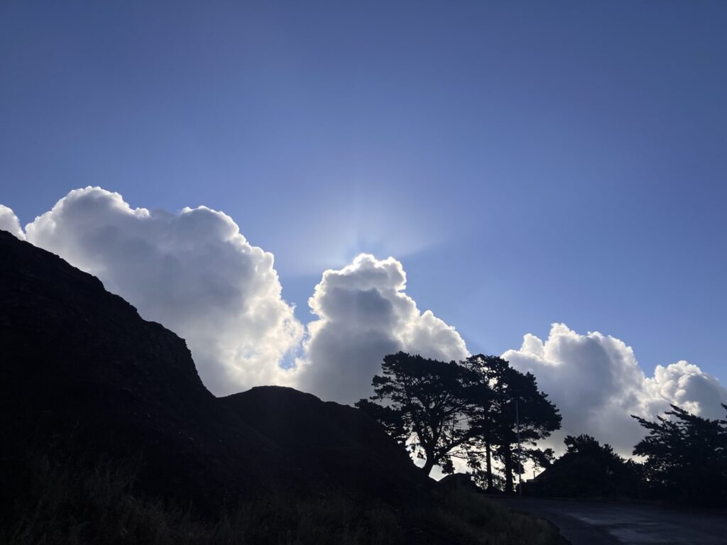 Silhouette of hill and trees, behind are big fluffy clouds against a blue sky. One cloud has a little ray of jesus beams coming out the top. 