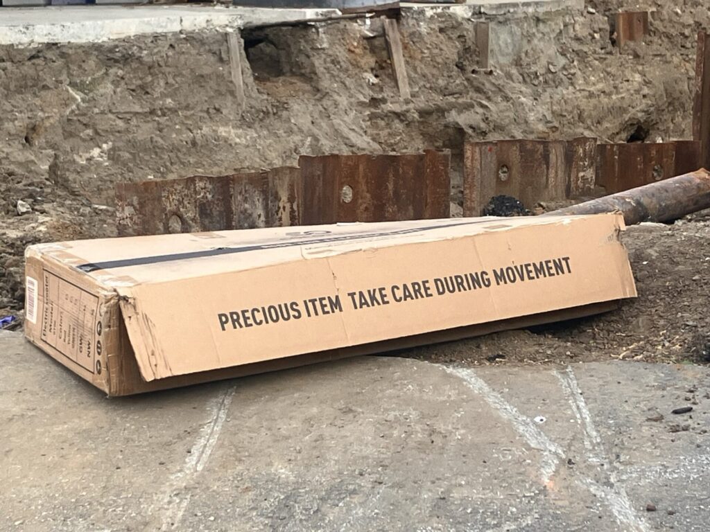 Oblong cardboard box with words printed on it: “PRECIOUS ITEM TAKE CARE DURING MOVEMENT”. Box sits on concrete next to a big pit. 